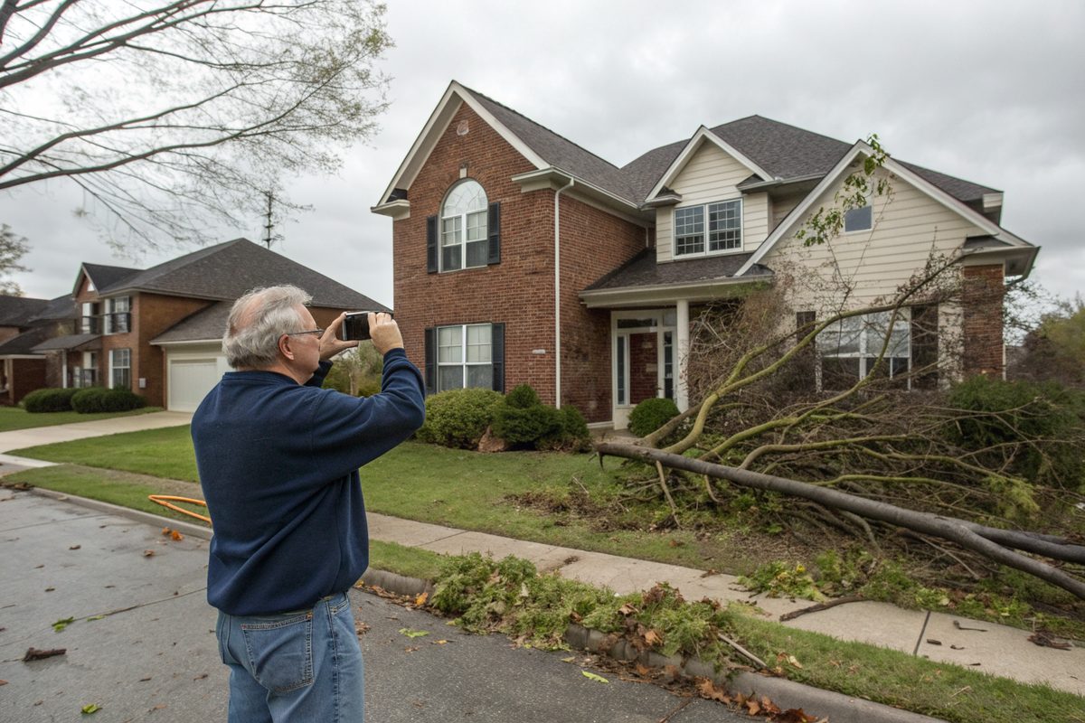 A homeowner photographing his house with a phone after a windstorm, with broken branches in the yard